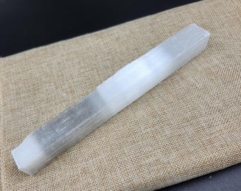 May include: A white selenite wand, rectangular in shape, rests on a tan fabric surface. The crystal is translucent with a layered appearance, and the ends are cut at an angle. The background is dark.