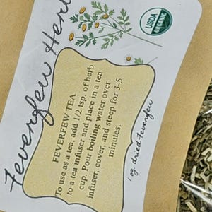 Puede incluir: Un paquete de t&eacute; de matricaria seca. La etiqueta incluye el texto "Feverfew Tea" e instrucciones de uso. La etiqueta tambi&eacute;n presenta un dibujo de una planta y el sello USDA Organic.