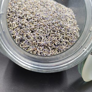 Puede incluir: Un tarro de cristal transparente lleno de capullos de lavanda secos. Los capullos de lavanda son una mezcla de morado y verde, llenando el tarro. El tarro est&aacute; sobre una superficie oscura.