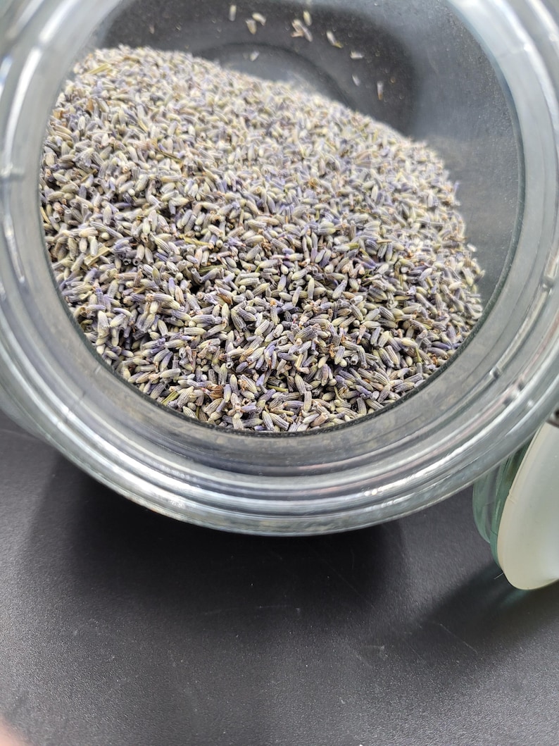 Puede incluir: Un tarro de cristal transparente lleno de capullos de lavanda secos. Los capullos de lavanda son una mezcla de morado y verde, llenando el tarro. El tarro est&aacute; sobre una superficie oscura.