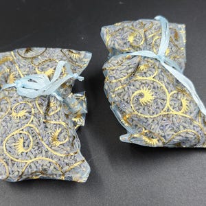 Puede incluir: Dos peque&ntilde;os saquitos llenos de lavanda seca. Los saquitos est&aacute;n hechos de una tela azul claro transparente con dise&ntilde;os dorados. Cada uno est&aacute; atado con una cinta azul claro. La lavanda es de color p&uacute;rpura p&aacute;lido.