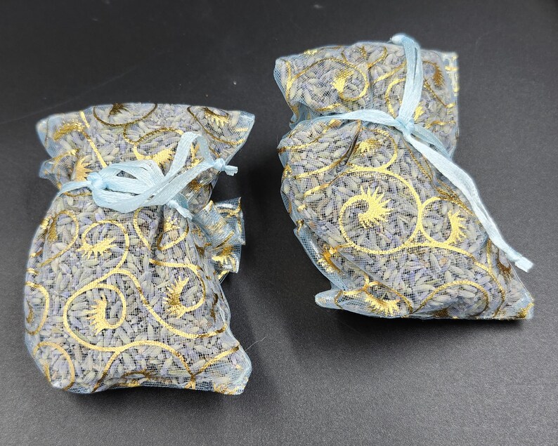 Puede incluir: Dos peque&ntilde;os saquitos llenos de lavanda seca. Los saquitos est&aacute;n hechos de una tela azul claro transparente con dise&ntilde;os dorados. Cada uno est&aacute; atado con una cinta azul claro. La lavanda es de color p&uacute;rpura p&aacute;lido.
