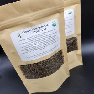 May include: Two brown paper bags filled with dried Krishna Holy Basil Leaf (Tulsi). Each bag has a white label with text, including "USDA Organic" and "1 oz.". The bags are sealed and sit against a dark background.