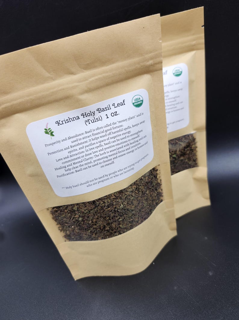 May include: Two brown paper bags filled with dried Krishna Holy Basil Leaf (Tulsi). Each bag has a white label with text, including "USDA Organic" and "1 oz.". The bags are sealed and sit against a dark background.