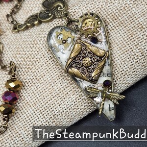 May include: Steampunk-style heart pendant with gears, a dragonfly, and a decorative square element. The pendant is primarily gold and silver, with accents of purple and white. The chain is also gold. The text "TheSteampunkBuddha" is at the bottom.