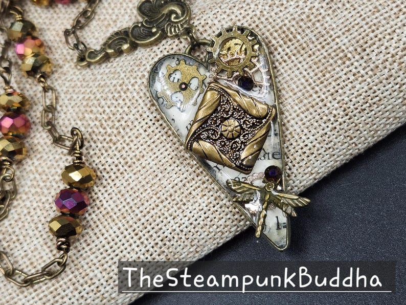 May include: Steampunk-style heart pendant with gears, a dragonfly, and a decorative square element. The pendant is primarily gold and silver, with accents of purple and white. The chain is also gold. The text "TheSteampunkBuddha" is at the bottom.