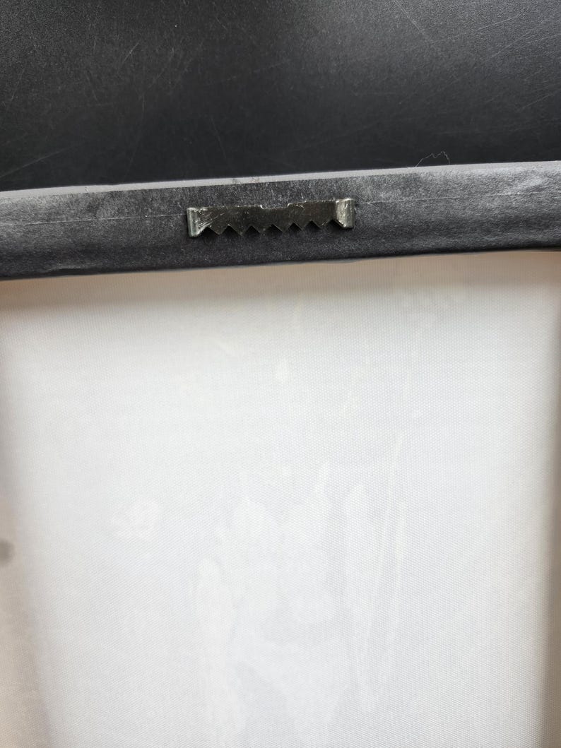 Puede incluir: Primer plano de la parte posterior de una impresi&oacute;n en lienzo enmarcada. El marco negro tiene un colgador de dientes de sierra para montaje en pared. El lienzo es blanco con un sutil patr&oacute;n floral.