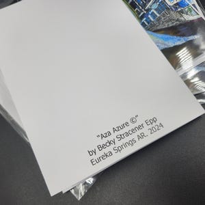 Op de afbeelding: Een stapel witte kunstdrukken met de tekst "Aza Azure &copy;" van Becky Stracener Epp, Eureka Springs AR. 2024. De hoek van een kleurrijke kunstdruk is zichtbaar in een doorzichtige plastic hoes.