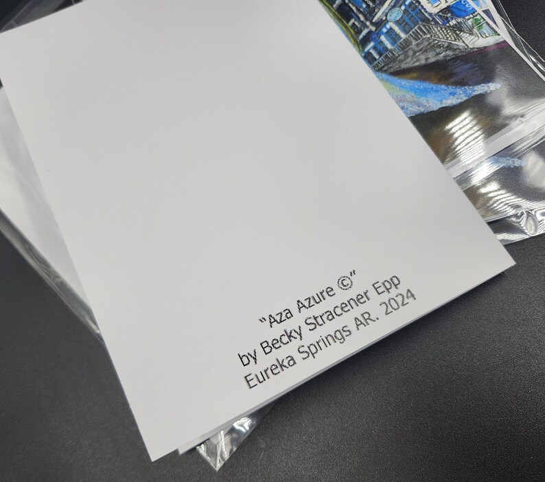 Op de afbeelding: Een stapel witte kunstdrukken met de tekst "Aza Azure &copy;" van Becky Stracener Epp, Eureka Springs AR. 2024. De hoek van een kleurrijke kunstdruk is zichtbaar in een doorzichtige plastic hoes.