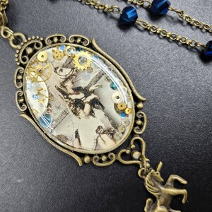 May include: An antique brass-toned pendant necklace with an oval-shaped centerpiece. The centerpiece features a vintage illustration with gears and blue and gold accents. A small brass unicorn charm hangs below the pendant. The necklace chain has blue beads.