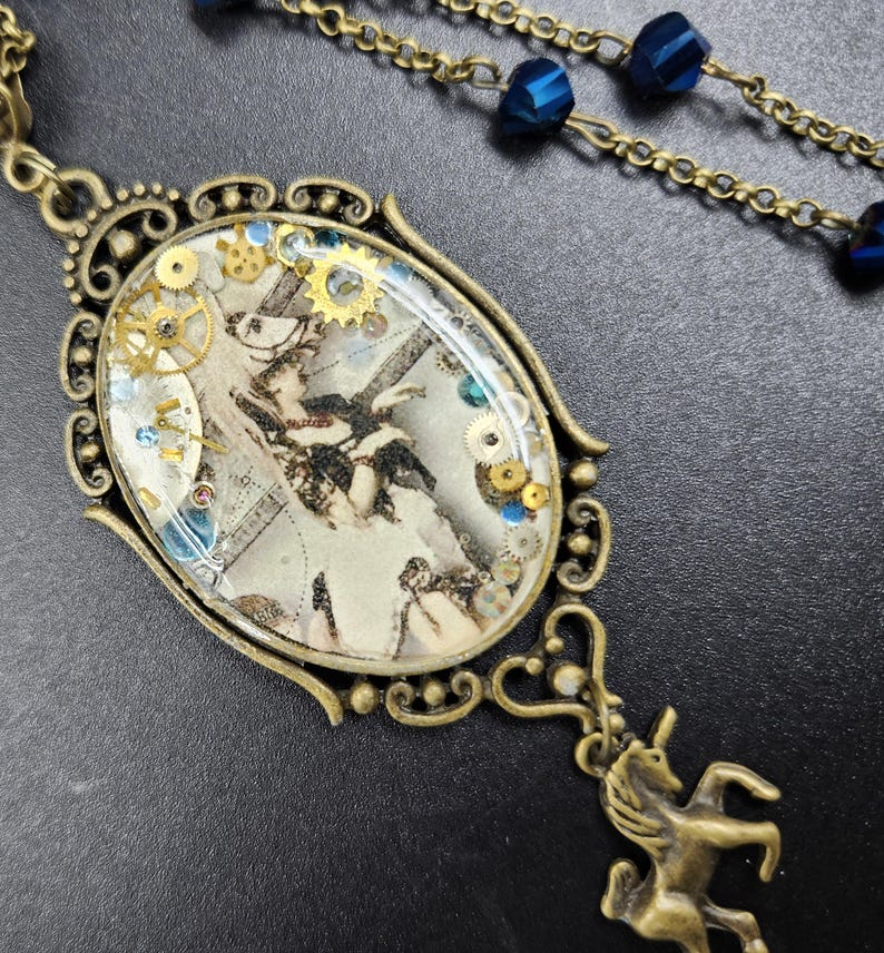 May include: An antique brass-toned pendant necklace with an oval-shaped centerpiece. The centerpiece features a vintage illustration with gears and blue and gold accents. A small brass unicorn charm hangs below the pendant. The necklace chain has blue beads.