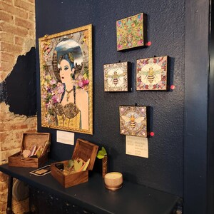 May include: A collection of wall art featuring a large framed portrait of a person with blue hair surrounded by flowers, alongside four smaller square canvases depicting bees. Two wooden boxes and a small candle sit on a shelf below.
