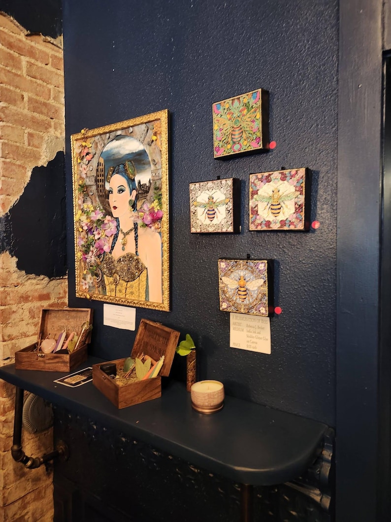 May include: A collection of wall art featuring a large framed portrait of a person with blue hair surrounded by flowers, alongside four smaller square canvases depicting bees. Two wooden boxes and a small candle sit on a shelf below.