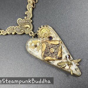 May include: Steampunk-style heart pendant necklace with intricate brass gears, filigree, and a dragonfly charm. The pendant features a square, ornate design and a small purple gem. The necklace chain and decorative elements are gold-toned.