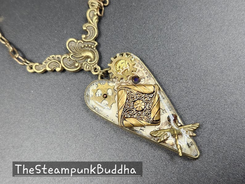 May include: Steampunk-style heart pendant necklace with intricate brass gears, filigree, and a dragonfly charm. The pendant features a square, ornate design and a small purple gem. The necklace chain and decorative elements are gold-toned.