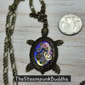 K&ouml;nnte beinhalten: Eine bronzefarbene Schildkr&ouml;ten-Halskette mit einem violetten ovalen Inlay, das goldfarbene Uhrwerk-Zahnr&auml;der und florale Muster enth&auml;lt. Der Anh&auml;nger wird neben einer US-Vierteldollarm&uuml;nze zum Gr&ouml;&szlig;envergleich gezeigt und h&auml;ngt an einer passenden Bronzekette.