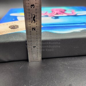 May include: A canvas art piece featuring a blue ocean scene with a pink octopus on a surfboard. The canvas is approximately 1 inch thick, measured with a ruler. A small wooden tag is visible on the left side.