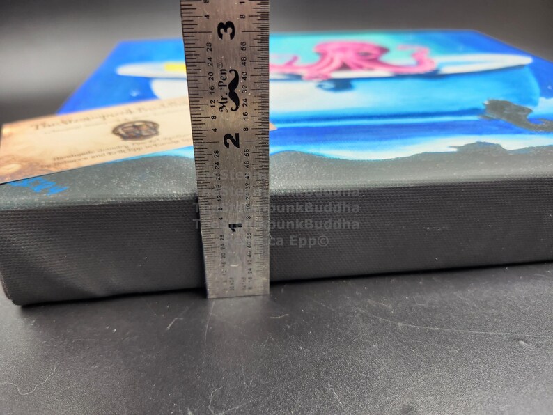 May include: A canvas art piece featuring a blue ocean scene with a pink octopus on a surfboard. The canvas is approximately 1 inch thick, measured with a ruler. A small wooden tag is visible on the left side.
