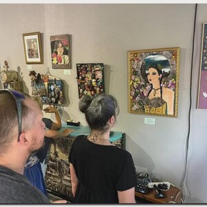 May include: A gallery wall featuring multiple framed artworks, including a central portrait of a person in a gold frame surrounded by floral elements, alongside smaller framed pieces and a mixed-media assemblage on a light-colored wall.
