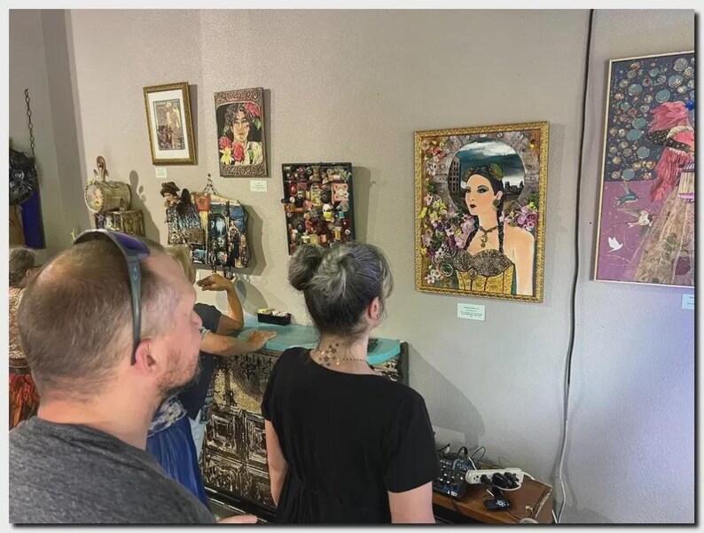 May include: A gallery wall featuring multiple framed artworks, including a central portrait of a person in a gold frame surrounded by floral elements, alongside smaller framed pieces and a mixed-media assemblage on a light-colored wall.