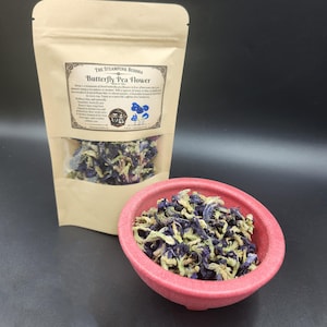 May include: A tan stand-up pouch labeled The Steampunk Buddha Butterfly Pea Flower containing dried blue and cream-colored flowers, accompanied by a small, round, textured red bowl filled with the same dried butterfly pea flowers.