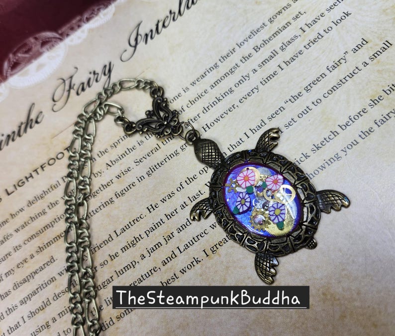 K&ouml;nnte beinhalten: Eine schildkr&ouml;tenf&ouml;rmige Anh&auml;ngerkette in Bronzefarbe mit einer Metallkette und einem Schmetterlingsverbindungsst&uuml;ck. Der Panzer enth&auml;lt eine violette Harzeinlage, die mit kleinen bunten Blumenformen und Metallzahnr&auml;dern verziert ist. Der Text TheSteampunkBuddha ist sichtbar.