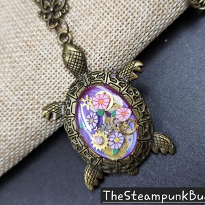 K&ouml;nnte beinhalten: Eine bronzefarbene Schildkr&ouml;ten-Anh&auml;nger-Halskette mit einer violetten ovalen Einlage. Die Einlage enth&auml;lt kleine goldfarbene Zahnr&auml;der, wei&szlig;-violette blumenf&ouml;rmige Anh&auml;nger und gr&uuml;ne Blattakzente. Der Text TheSteampunkBuddha ist unten zu sehen.