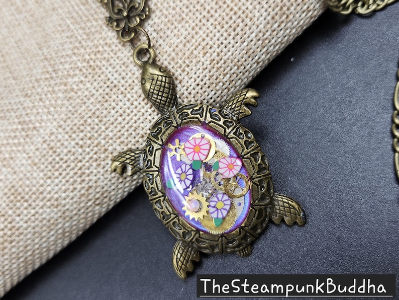 K&ouml;nnte beinhalten: Eine bronzefarbene Schildkr&ouml;ten-Anh&auml;nger-Halskette mit einer violetten ovalen Einlage. Die Einlage enth&auml;lt kleine goldfarbene Zahnr&auml;der, wei&szlig;-violette blumenf&ouml;rmige Anh&auml;nger und gr&uuml;ne Blattakzente. Der Text TheSteampunkBuddha ist unten zu sehen.
