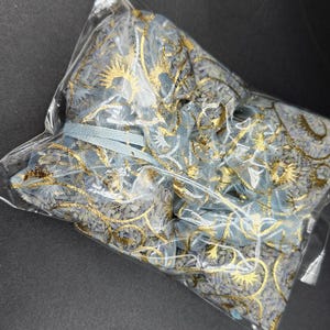 Puede incluir: Una bolsa de pl&aacute;stico transparente que contiene varias bolsas de tela de malla azul y dorada. Las bolsas est&aacute;n decoradas con patrones florales dorados y atadas con una cinta azul claro. La est&eacute;tica general es delicada y ornamentada.