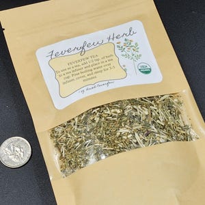 Puede incluir: Una bolsa de papel marr&oacute;n con una ventana transparente que contiene hierba de matricaria seca. La bolsa tiene una etiqueta con el texto "Feverfew Herb" e instrucciones de uso. Una moneda est&aacute; al lado.