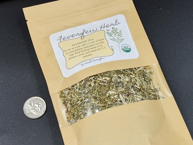 Puede incluir: Una bolsa de papel marr&oacute;n con una ventana transparente que contiene hierba de matricaria seca. La bolsa tiene una etiqueta con el texto "Feverfew Herb" e instrucciones de uso. Una moneda est&aacute; al lado.