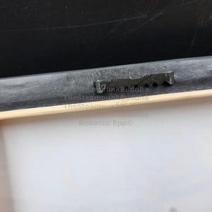 May include: Close-up of the back of a framed canvas print. The wooden frame is visible, along with a black backing and a metal sawtooth hanger. The canvas is white.