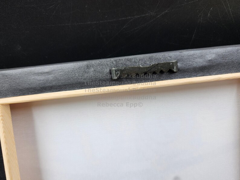 May include: Close-up of the back of a framed canvas print. The wooden frame is visible, along with a black backing and a metal sawtooth hanger. The canvas is white.