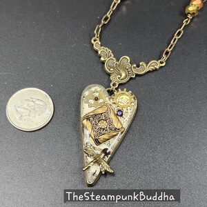 May include: A steampunk-style heart pendant necklace with a brass chain. The heart-shaped pendant features gears, a dragonfly, and a decorative square element. The text "TheSteampunkBuddha" is at the bottom.