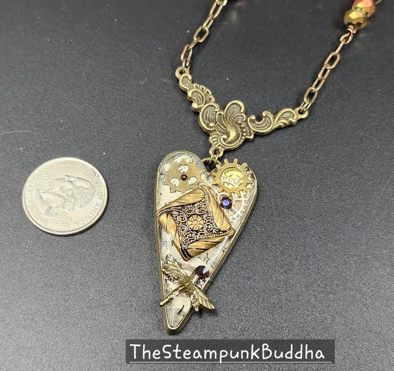 May include: A steampunk-style heart pendant necklace with a brass chain. The heart-shaped pendant features gears, a dragonfly, and a decorative square element. The text "TheSteampunkBuddha" is at the bottom.