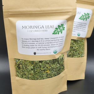 Op de afbeelding: Drie bruine papieren zakken gevuld met gedroogde Moringa-bladeren. Elke zak heeft een wit etiket met de tekst "MORINGA LEAF 1 OZ DRIED HERB" en instructies voor het zetten van thee. De zakken zijn USDA biologisch gecertificeerd.