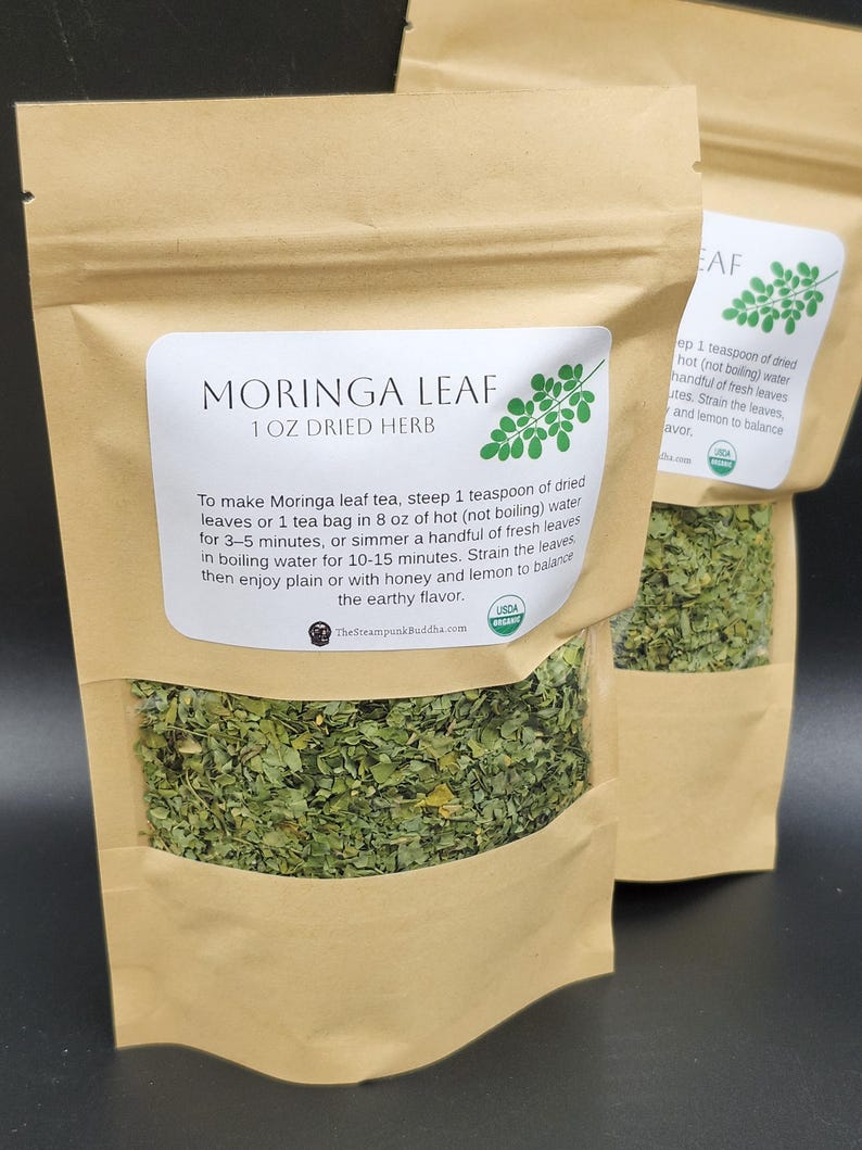 Op de afbeelding: Drie bruine papieren zakken gevuld met gedroogde Moringa-bladeren. Elke zak heeft een wit etiket met de tekst "MORINGA LEAF 1 OZ DRIED HERB" en instructies voor het zetten van thee. De zakken zijn USDA biologisch gecertificeerd.