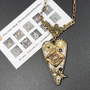 May include: A steampunk-style heart pendant necklace with a brass chain. The heart-shaped pendant features gears, a dragonfly, and a map design. The necklace has a vintage aesthetic and is likely handmade.
