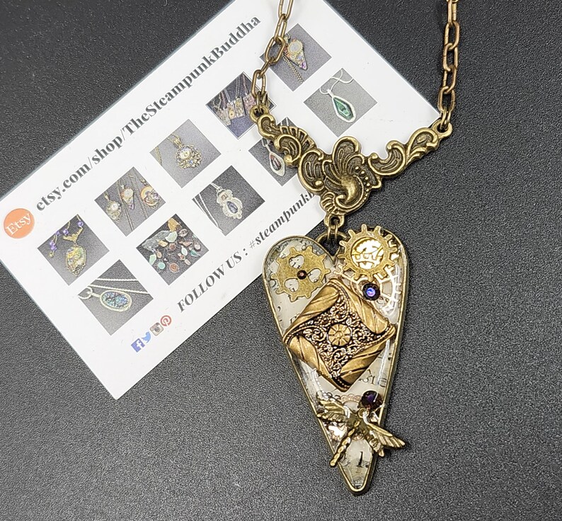 May include: A steampunk-style heart pendant necklace with a brass chain. The heart-shaped pendant features gears, a dragonfly, and a map design. The necklace has a vintage aesthetic and is likely handmade.