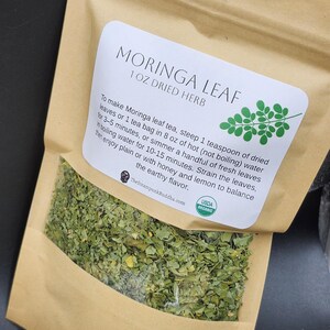 Op de afbeelding: Een bruine papieren zak met gedroogde Moringa bladthee. Het etiket luidt "MORINGA LEAF 1 OZ DRIED HERB" met instructies en een USDA Organic zegel. De zak bevat groene gedroogde bladeren.