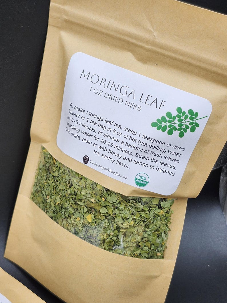 Op de afbeelding: Een bruine papieren zak met gedroogde Moringa bladthee. Het etiket luidt "MORINGA LEAF 1 OZ DRIED HERB" met instructies en een USDA Organic zegel. De zak bevat groene gedroogde bladeren.