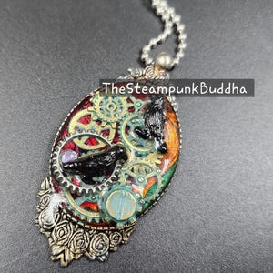 Peut inclure: Pendentif ovale avec un design steampunk. Le pendentif pr&eacute;sente un corbeau noir, des engrenages et une gemme violette sur un fond rouge, vert et or. Le texte "TheSteampunkBuddha" est visible sur le pendentif.