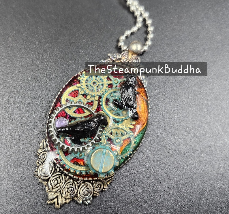 Peut inclure: Pendentif ovale avec un design steampunk. Le pendentif pr&eacute;sente un corbeau noir, des engrenages et une gemme violette sur un fond rouge, vert et or. Le texte "TheSteampunkBuddha" est visible sur le pendentif.