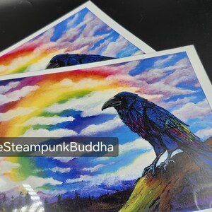 Op de afbeelding: Twee kunstprints met een raaf met blauwe en paarse veren, tegen een achtergrond van een regenboog en een bewolkte lucht. De raaf zit op een rots. De tekst "TheSteampunkBuddha" is zichtbaar linksonder op de print.