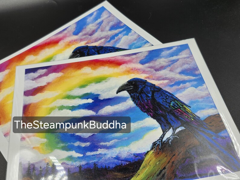 Op de afbeelding: Twee kunstprints met een raaf met blauwe en paarse veren, tegen een achtergrond van een regenboog en een bewolkte lucht. De raaf zit op een rots. De tekst "TheSteampunkBuddha" is zichtbaar linksonder op de print.
