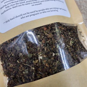 May include: A clear plastic bag filled with dried basil leaves, displayed on a brown paper background. The bag has a white label with text about the herb's properties. The text includes information about love, healing, and purification.