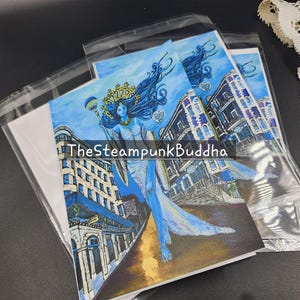 Op de afbeelding: Kunstafdrukken met een blauw getinte illustratie van een vrouw met lang, golvend haar, een kroon en een staf, tegen een achtergrond van gebouwen. De tekst "TheSteampunkBuddha" is zichtbaar. De afdrukken zijn verpakt in helder plastic.
