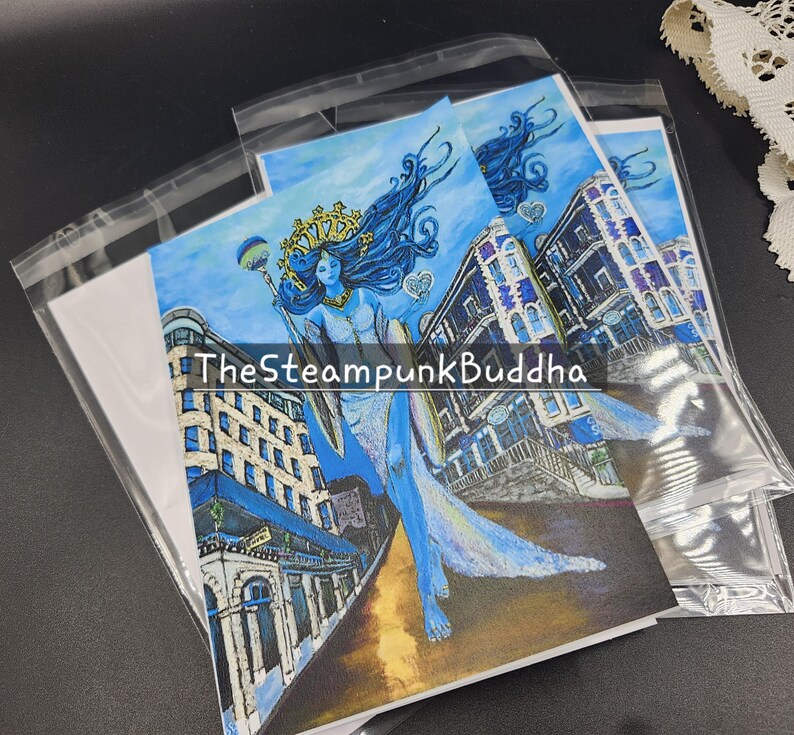 Op de afbeelding: Kunstafdrukken met een blauw getinte illustratie van een vrouw met lang, golvend haar, een kroon en een staf, tegen een achtergrond van gebouwen. De tekst "TheSteampunkBuddha" is zichtbaar. De afdrukken zijn verpakt in helder plastic.
