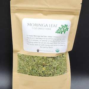 Op de afbeelding: Een bruine papieren zak met gedroogde Moringa-bladkruiden. Het etiket luidt "MORINGA LEAF 1 OZ DRIED HERB". Instructies voor het maken van thee staan op het etiket. De zak bevat groene gedroogde bladeren. USDA Organic gecertificeerd.