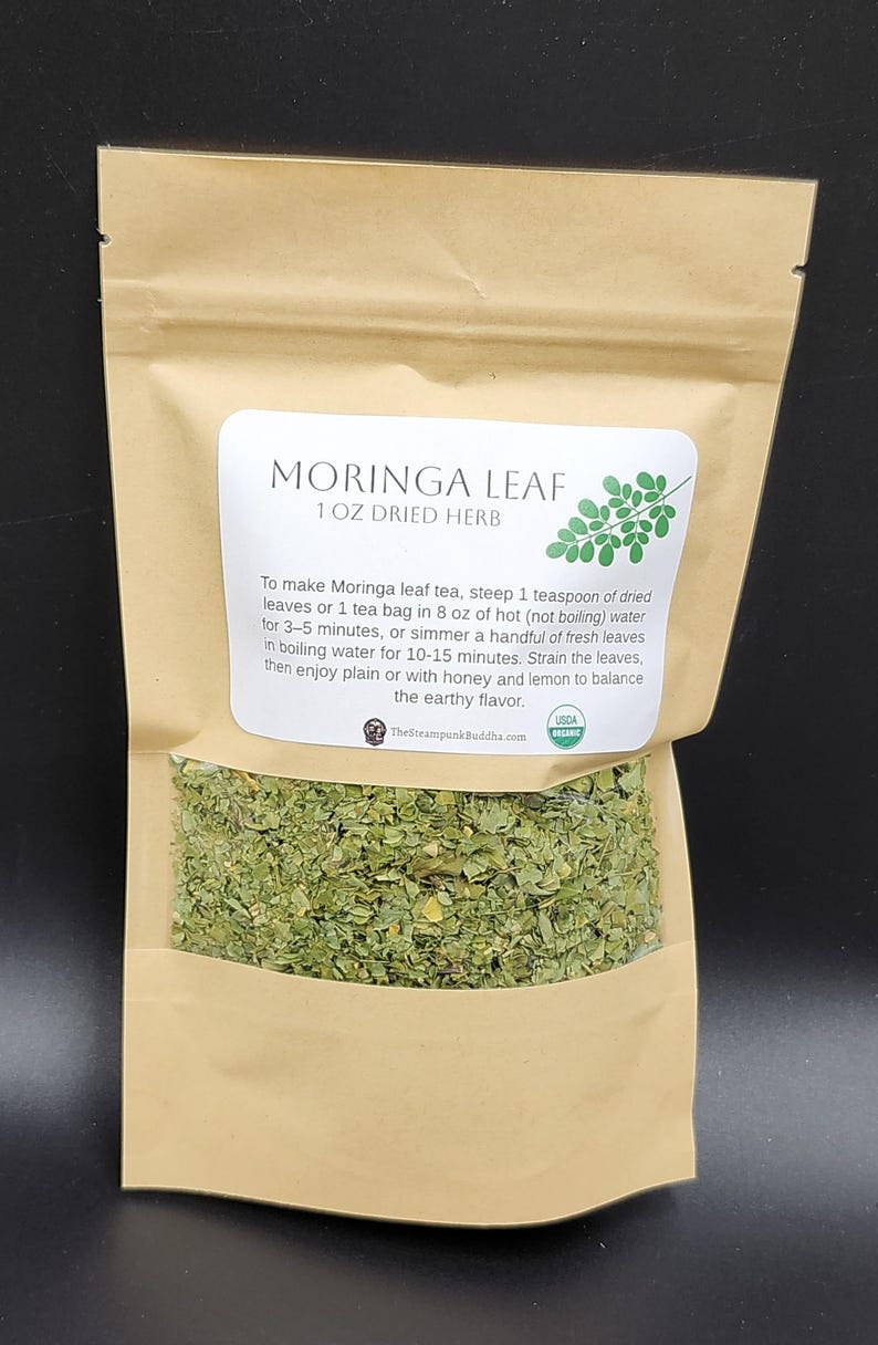 Op de afbeelding: Een bruine papieren zak met gedroogde Moringa-bladkruiden. Het etiket luidt "MORINGA LEAF 1 OZ DRIED HERB". Instructies voor het maken van thee staan op het etiket. De zak bevat groene gedroogde bladeren. USDA Organic gecertificeerd.